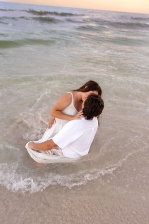 Romantic couple kissing in ocean waves at sunset, wearing white outfits on a beautiful beach.