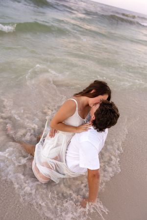 Romantic couple kissing in ocean waves at beach, woman in white dress, man in white shirt.