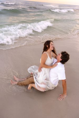 Couple laughing and sitting together in shallow beach waves at sunset, dressed in white.