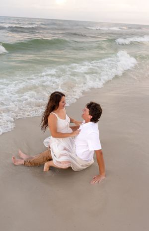 Romantic couple sitting together in wet sand at beach shoreline during golden sunset with ocean waves.
