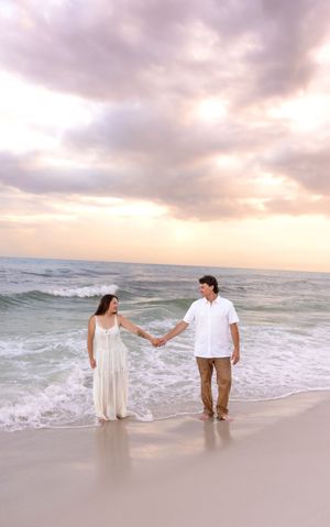 Couple holding hands on a sandy beach at sunset with gentle ocean waves behind them.
