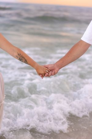Close-up of couple holding hands on beach, woman with floral tattoo wearing engagement ring by ocean waves.