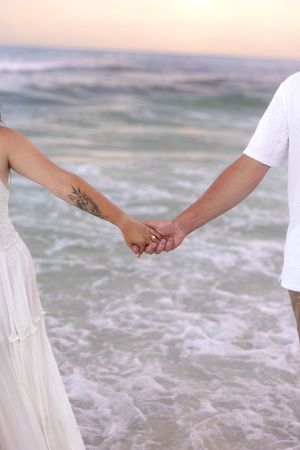 Couple holding hands on beach at sunset, woman with floral arm tattoo in white dress.