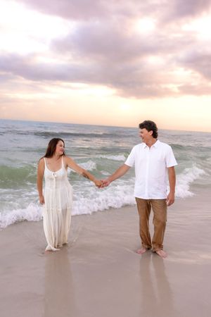 Couple holding hands and walking along sandy beach at sunset, woman in white dress, man in white shirt.