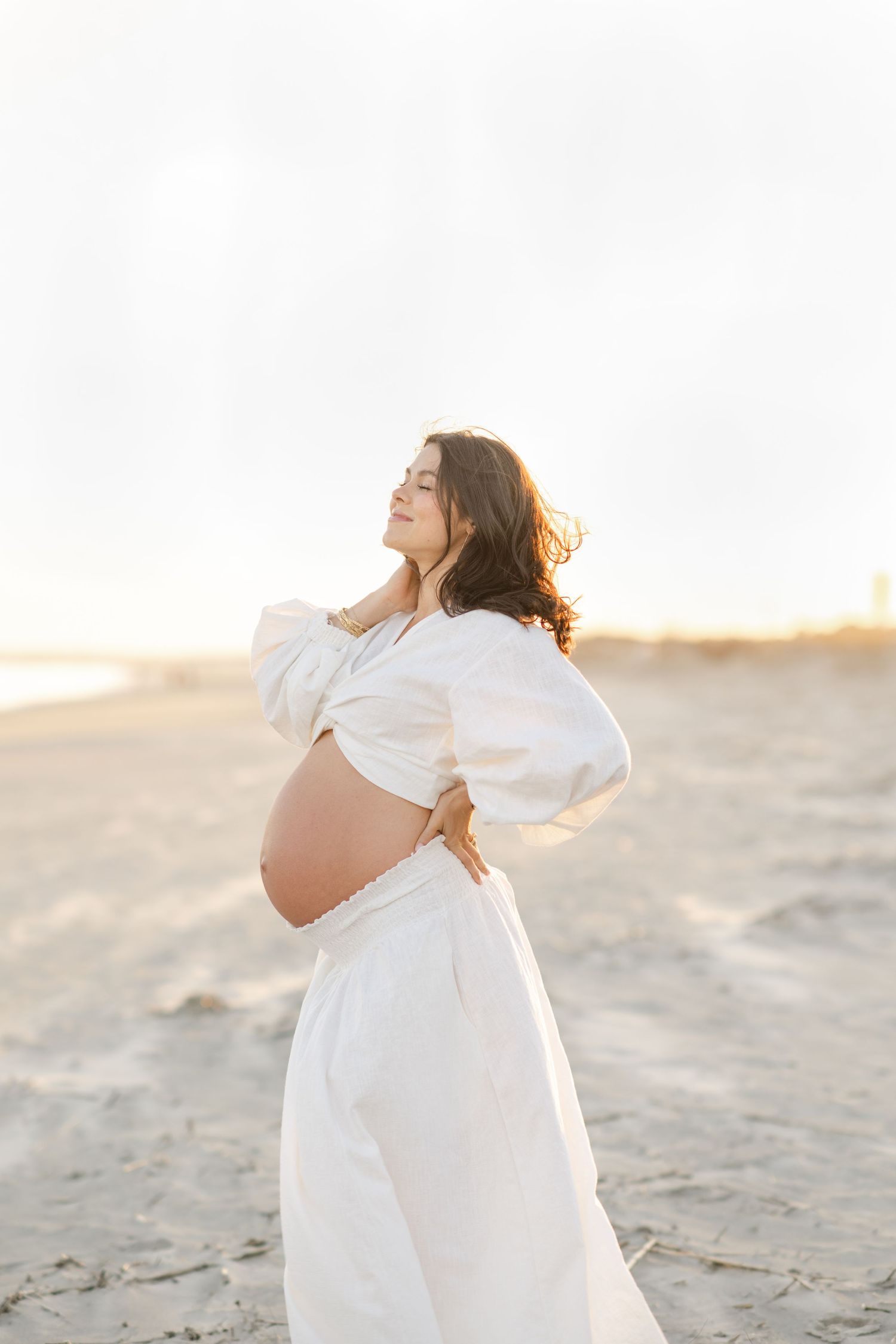 Pregnant woman in white crop top and skirt posing on sandy beach at golden hour sunset.