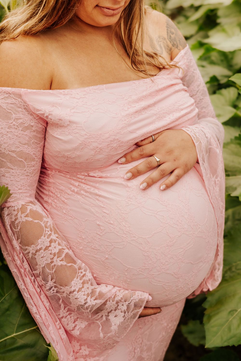 Pregnant woman in pink lace off-shoulder maternity gown cradling baby bump outdoors among green leaves.