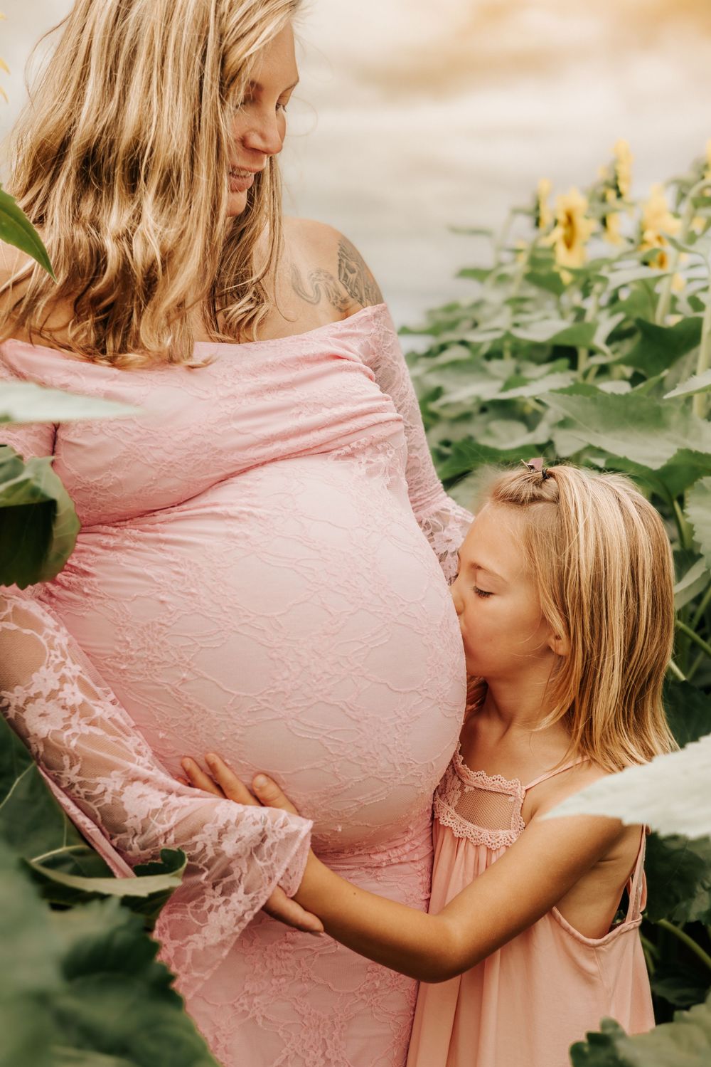 A young girl kisses her pregnant mother's belly outdoors among sunflowers, both wearing matching pink dresses.
