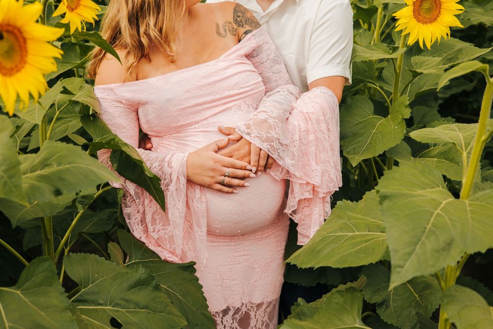 Pregnant woman in pink lace maternity dress with partner in sunflower field, cradling baby bump.