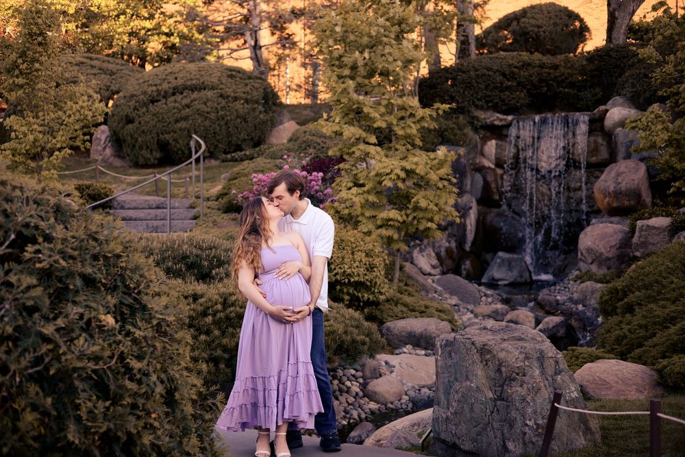 Expecting couple embraces tenderly in Japanese garden with waterfall, woman wearing lavender maternity dress.