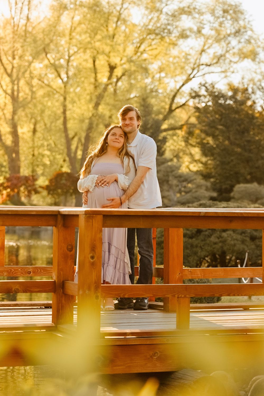 Couple poses on wooden bridge at golden hour, man embraces pregnant woman in pink dress surrounded by autumn trees.