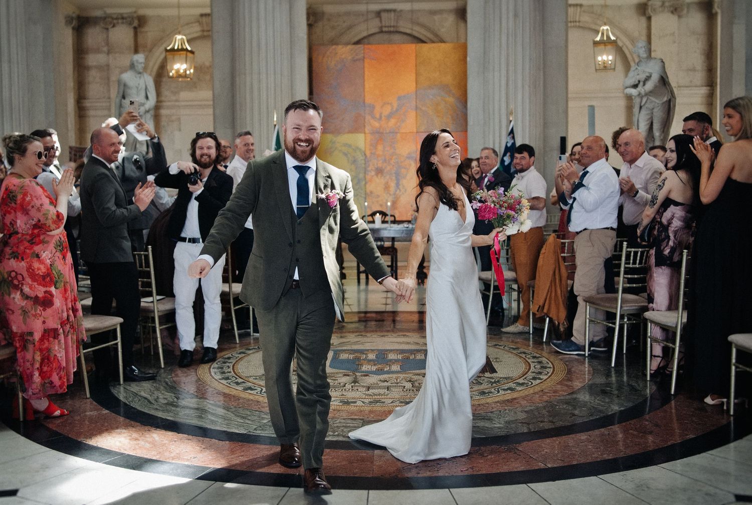 Joyful newlyweds walk down the aisle in an elegant marble hall, guests applauding their wedding ceremony.