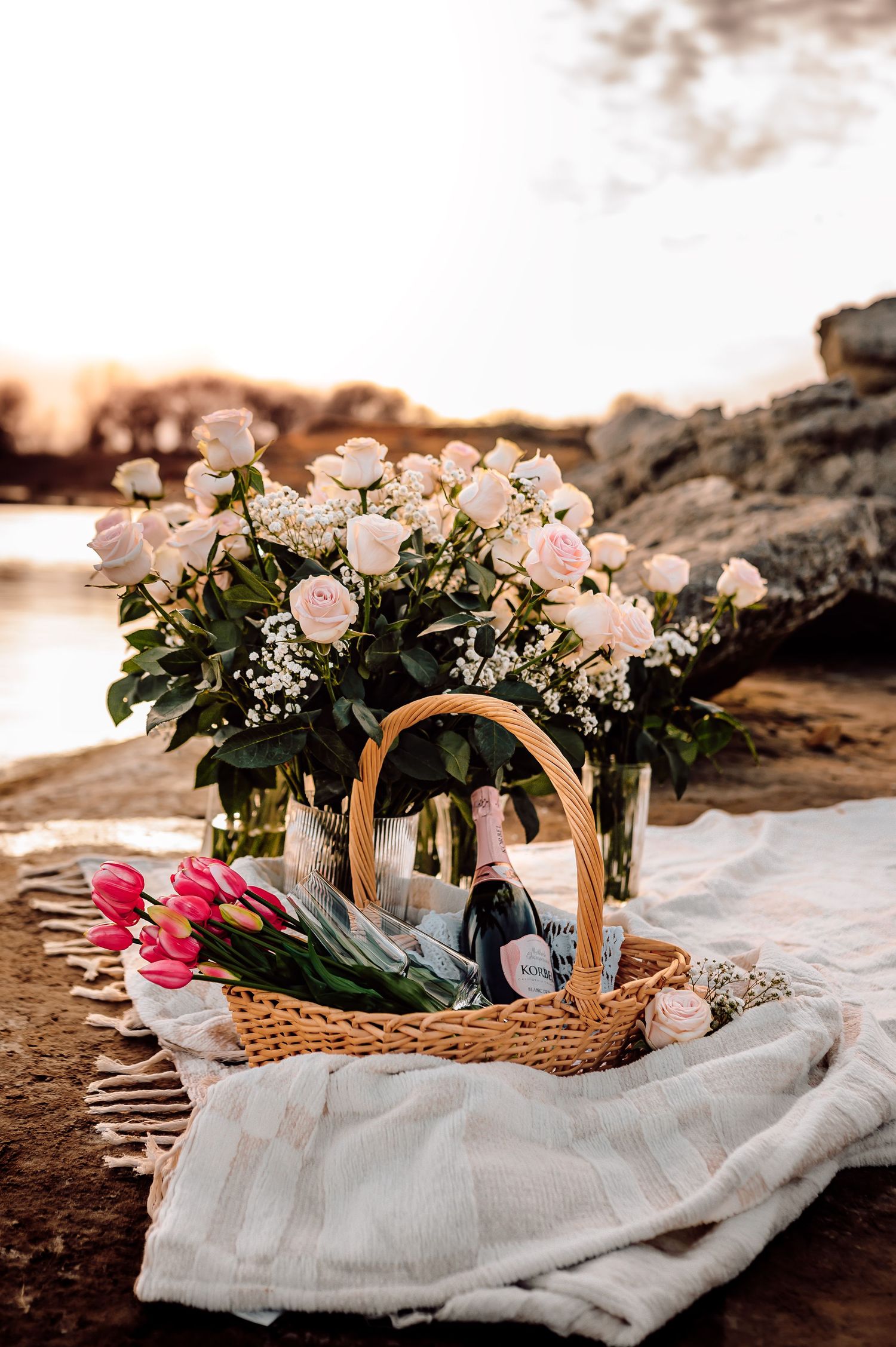 Romantic picnic setup with pink roses, tulips, champagne, and wicker basket at sunset.