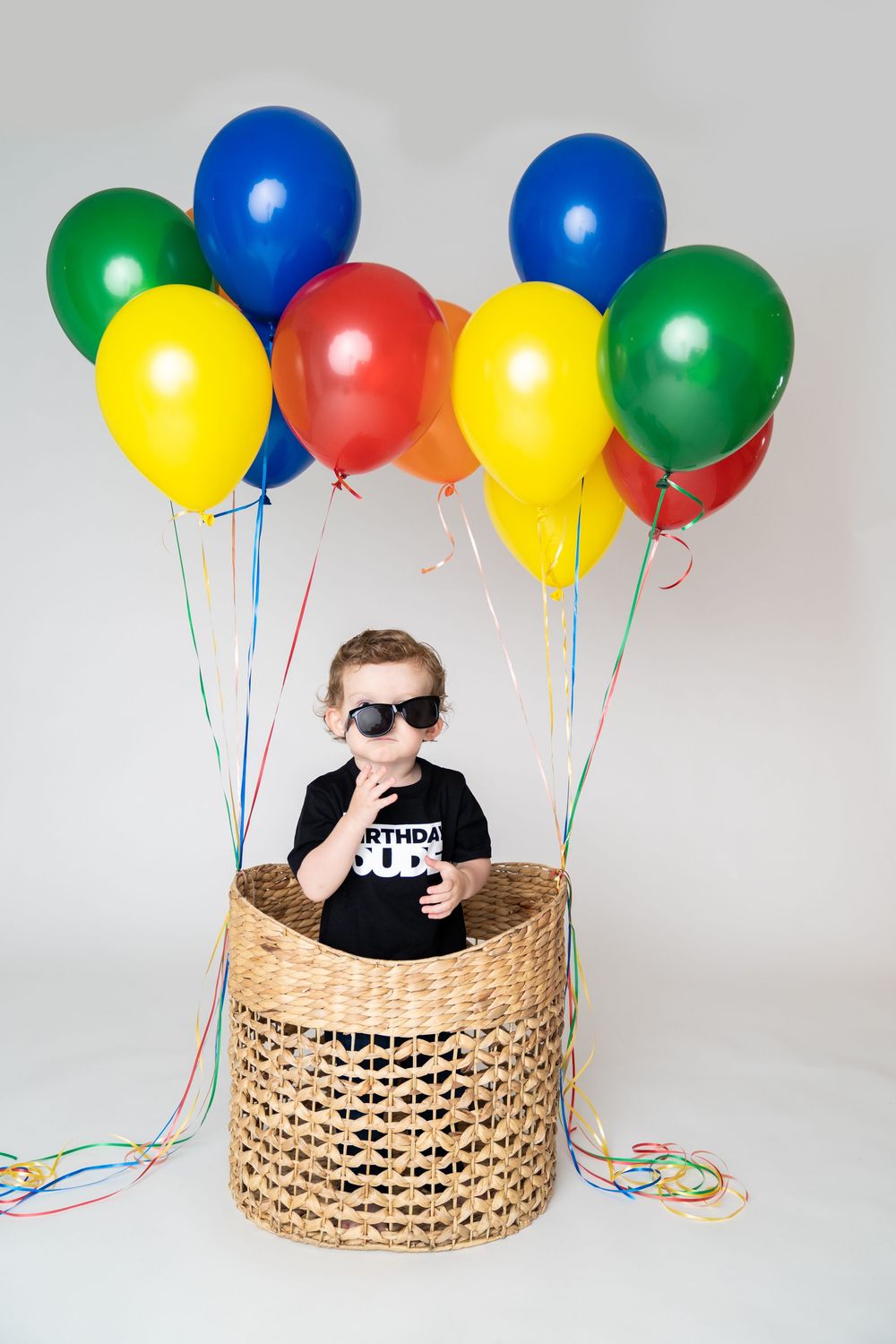 A young boy wearing sunglasses sits in a wicker basket with colorful balloons, celebrating his birthday.