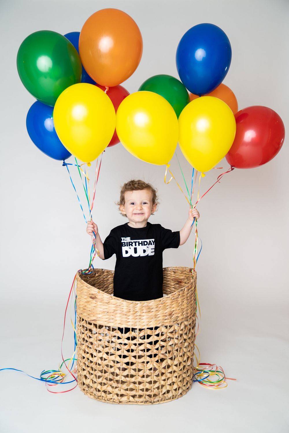 A smiling toddler in a Birthday Dude shirt stands in a wicker basket holding colorful balloons.