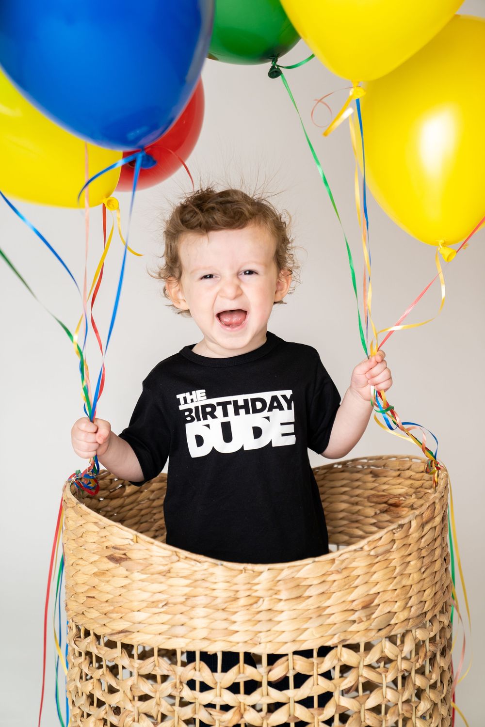 Happy toddler in a black Birthday Dude shirt sits in a wicker basket surrounded by colorful balloons.