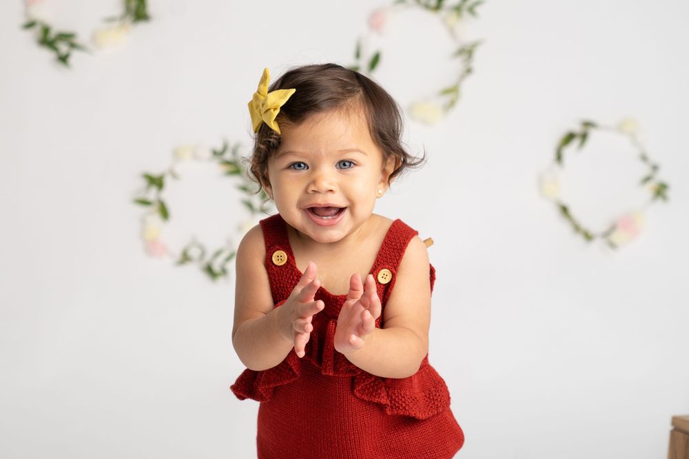 Smiling baby girl in red romper with yellow bow clapping hands against white floral wreath backdrop.