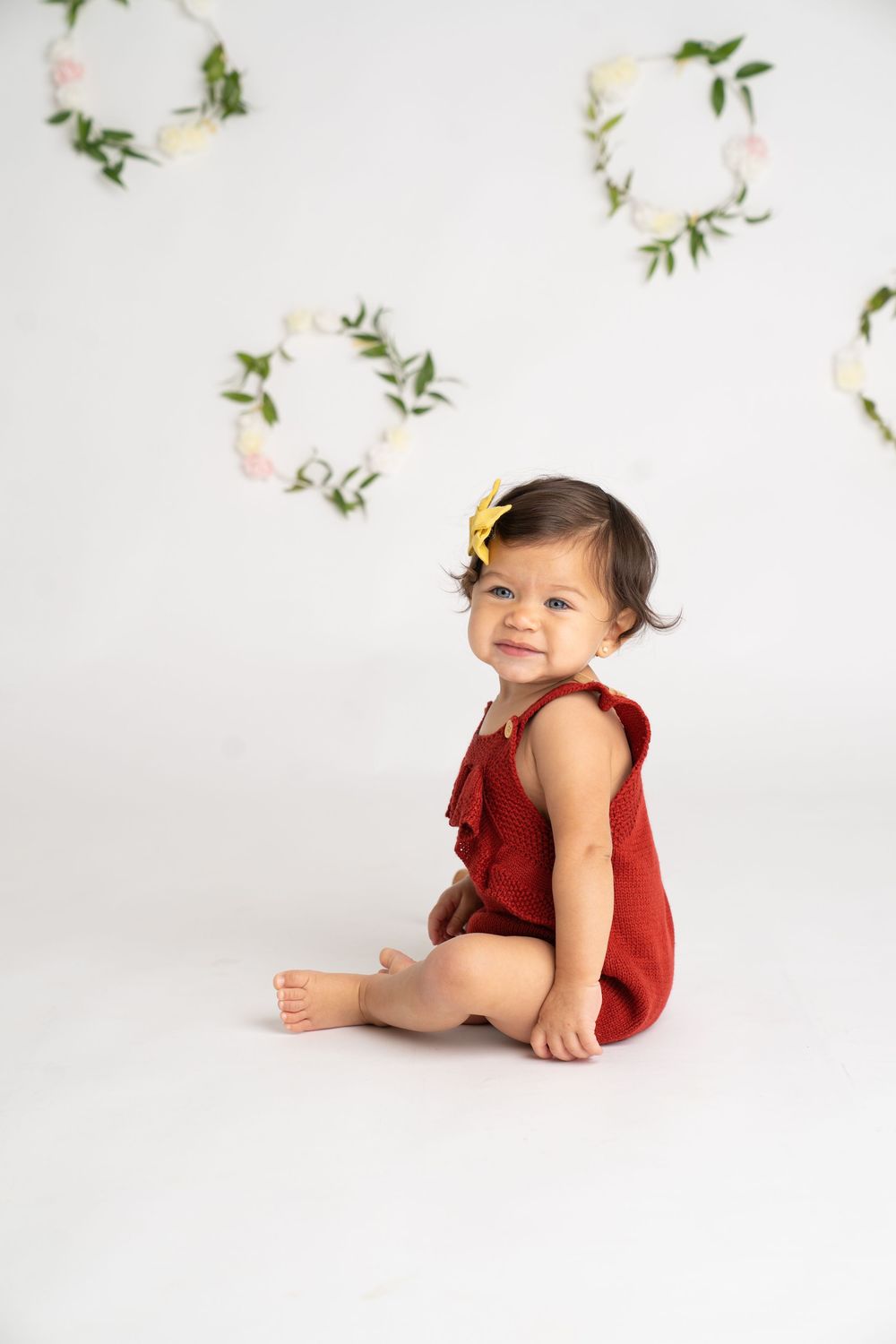 Smiling baby girl in red romper with yellow bow sits on white floor with floral wreath backdrop.