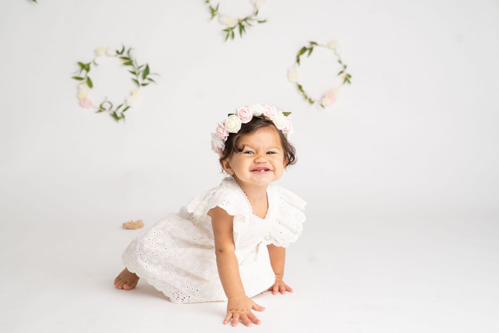 Happy baby girl in white dress and floral crown crawling on white floor with flower wreaths backdrop.