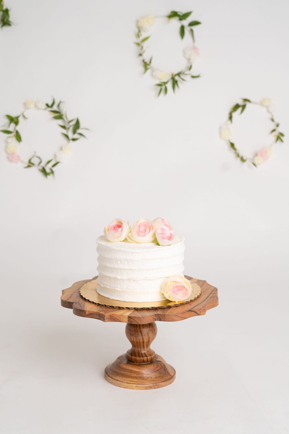 White frosted cake with pink roses on wooden stand, floral wreaths on white wall backdrop.