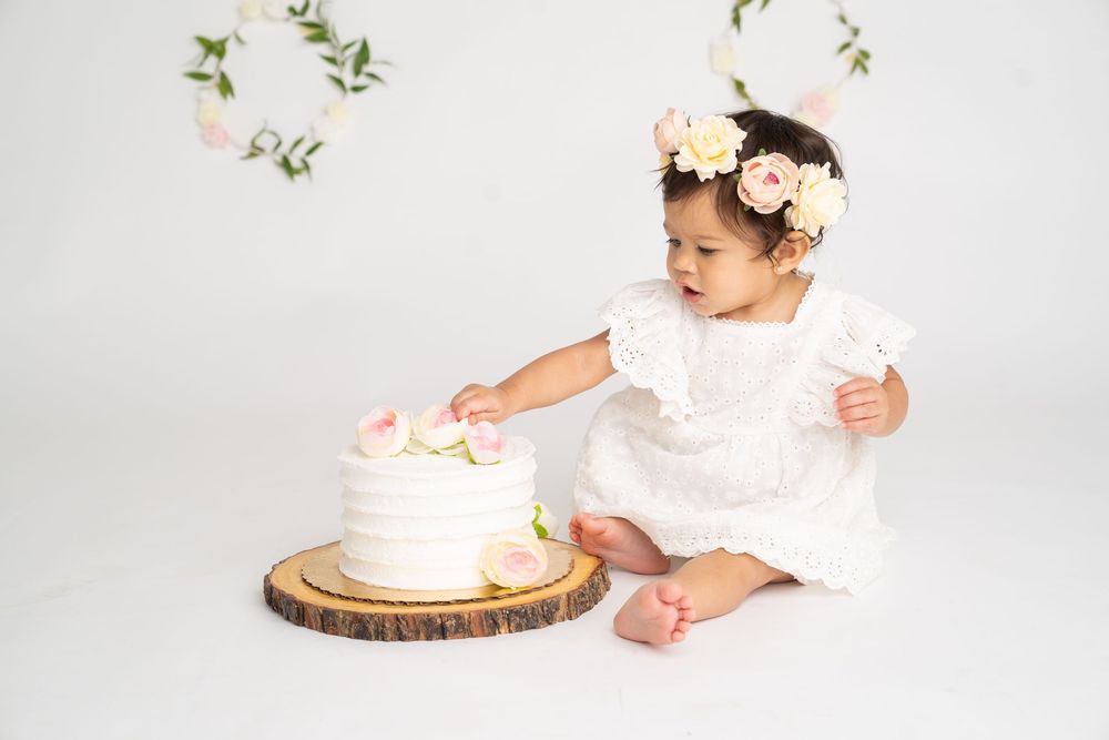 Baby girl in white dress and floral crown reaches for her first birthday cake on a wood slice.