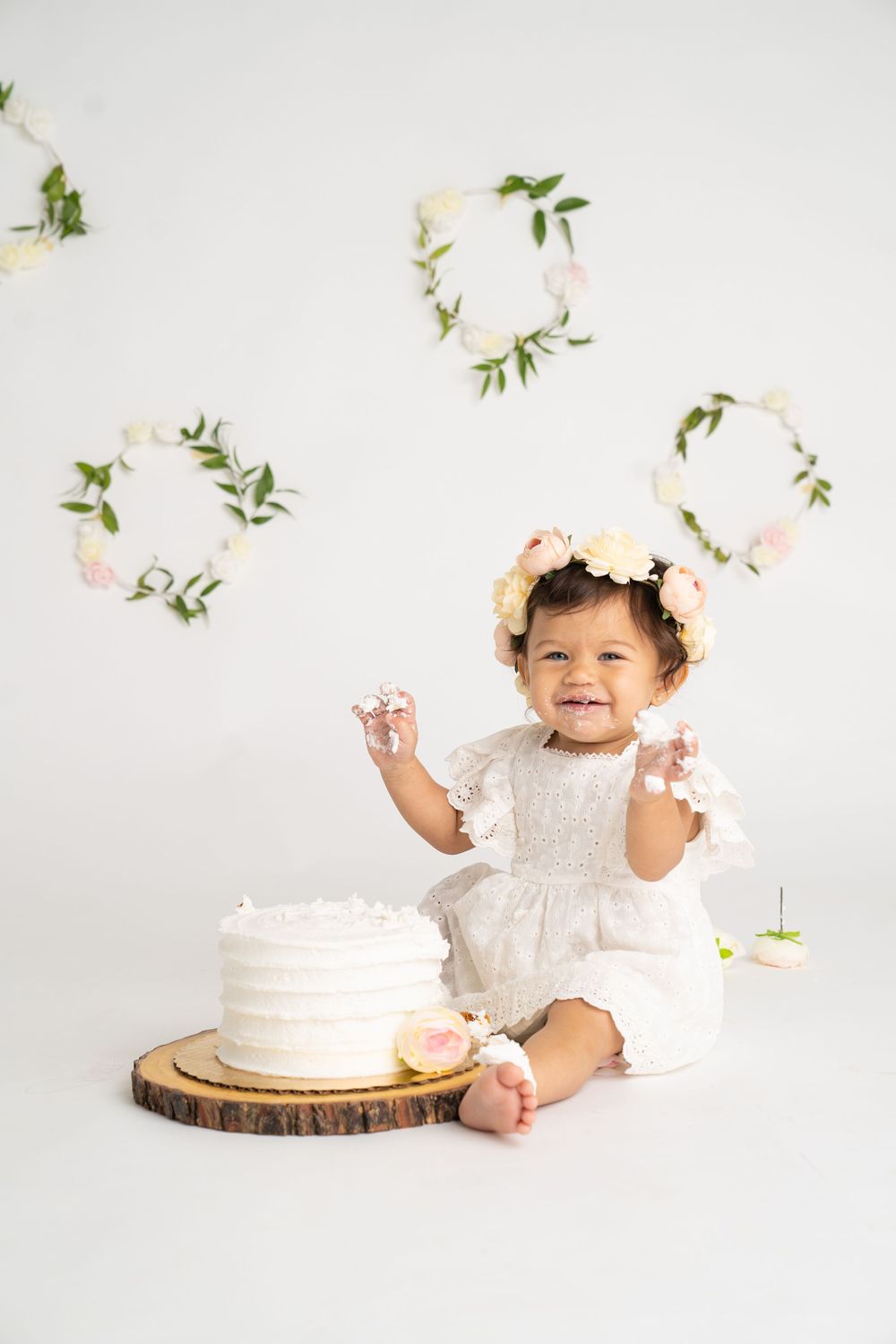 Happy baby girl in white dress with floral crown sitting beside first birthday smash cake on wood slice.
