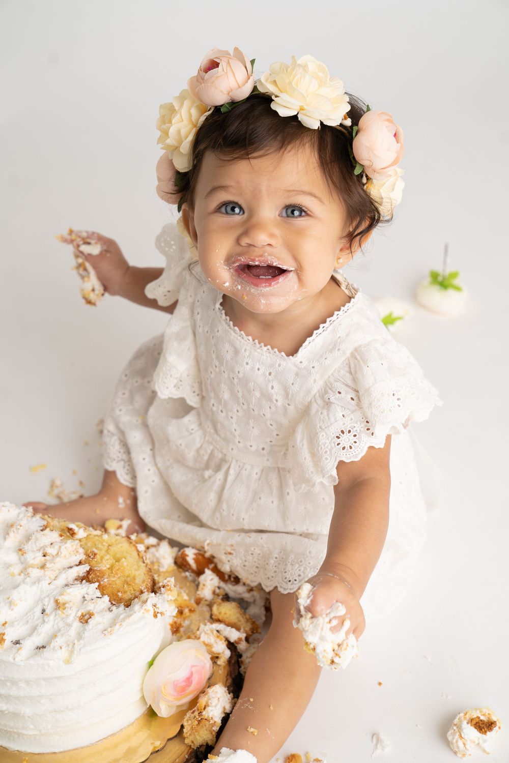 Baby girl in white dress and floral crown smiling during first birthday cake smash photo session.