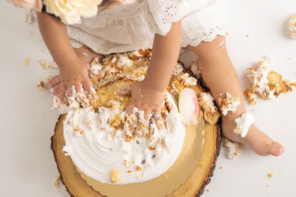 Baby girl in white dress smashing first birthday cake, hands covered in frosting during cake smash photo shoot.