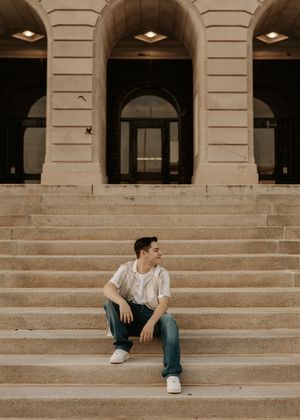 Young man in white t-shirt and jeans sitting on stone steps of a grand neoclassical building.
