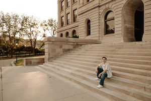 Person sitting on steps of a grand stone building at sunset, wearing white jacket and teal pants.