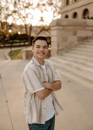 Smiling young man with crossed arms poses outdoors near stone building steps at golden hour.