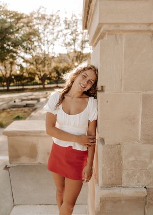 Smiling young woman in white crop top and red mini skirt leaning against a stone wall outdoors.