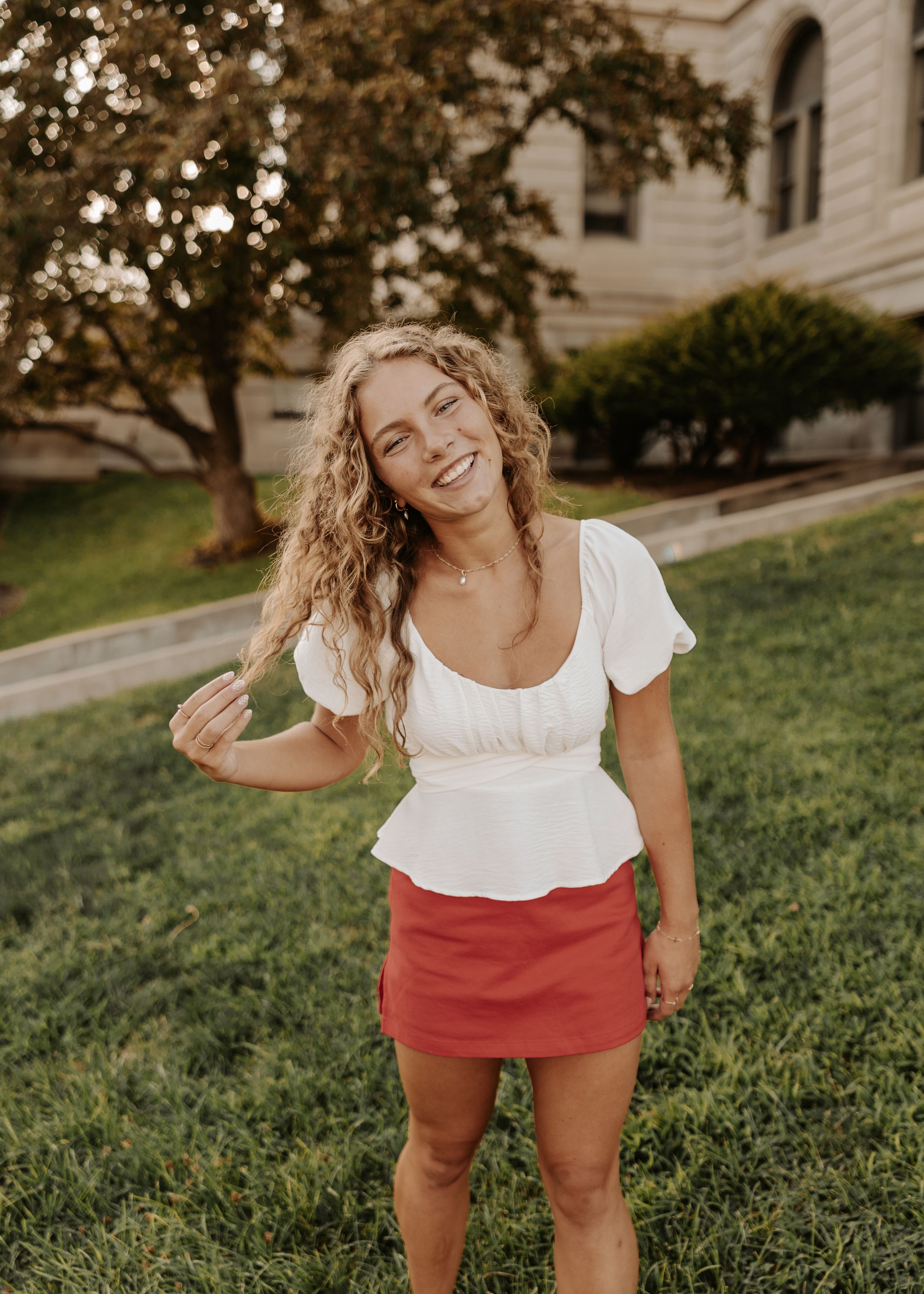 Smiling young woman with curly blonde hair wearing white peplum top and red mini skirt standing on green grass.