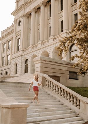 Young woman in white top and red shorts walks down grand stone steps of a neoclassical building.