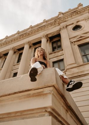 Young woman with curly hair sitting on a stone ledge of a classical building, wearing black top and white pants.
