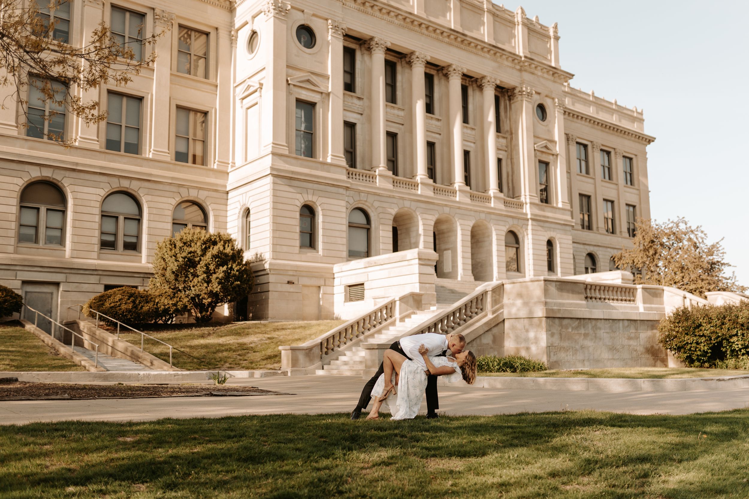 Couple sharing a romantic dip kiss on lawn in front of grand white historic building on sunny day.