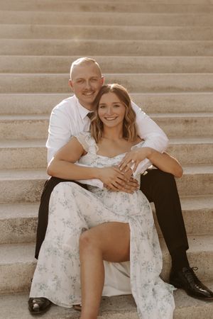 A smiling couple sits on stone steps, the man in white shirt embracing a woman in a floral dress during a golden hour engagement photo.