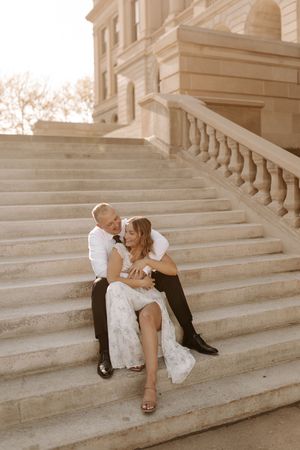 Couple in wedding attire sitting closely on grand stone steps of a classical building in warm golden light.