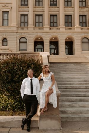 Couple poses on stone steps of elegant building, man in white shirt and tie, woman in floral white dress.