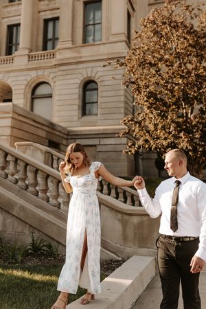 Couple in wedding attire holding hands walking outside a grand stone building with balustrade staircase.