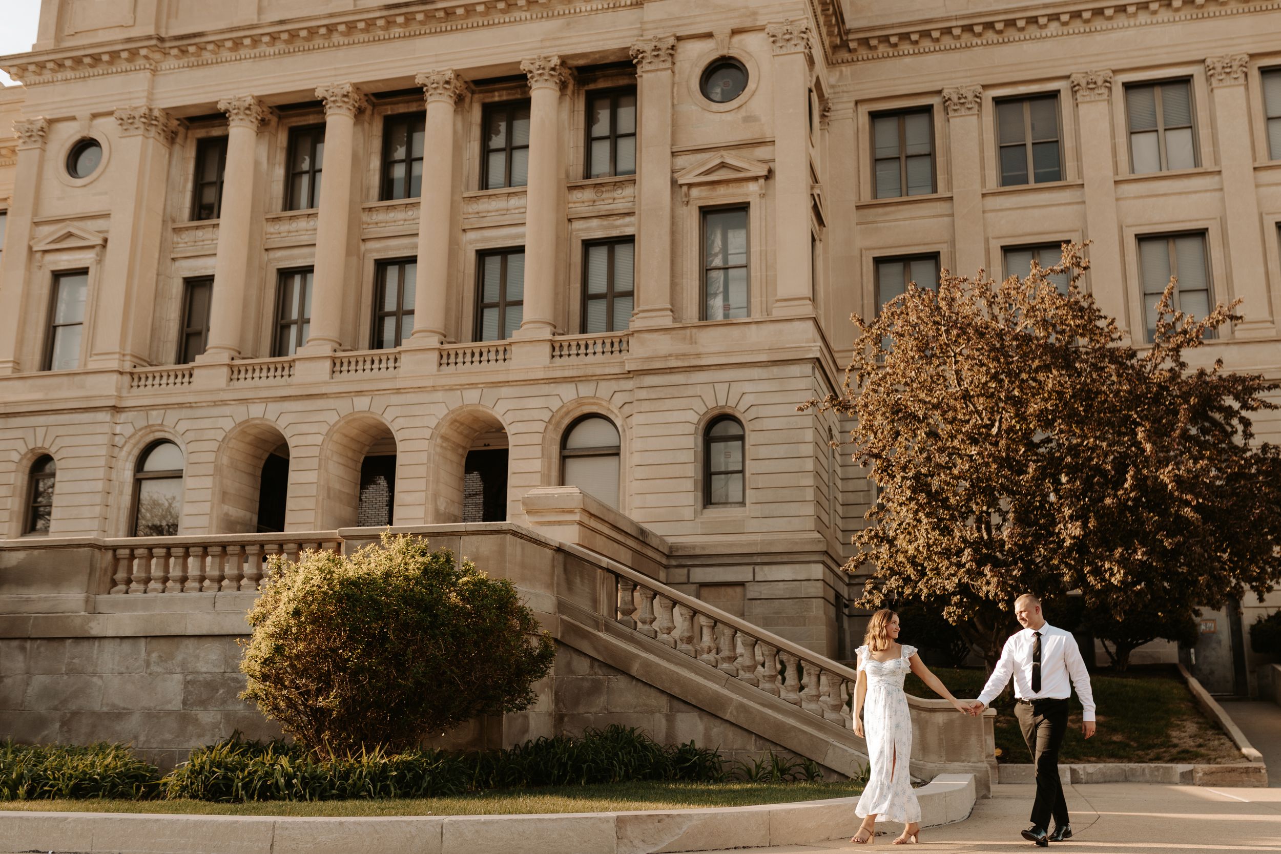 Bride and groom hold hands walking outside a grand neoclassical building with autumn trees in warm sunlight.
