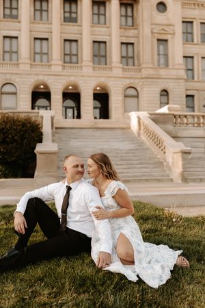A couple in wedding attire sits on grass in front of a grand stone building, sharing a tender romantic moment.