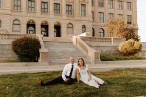 Couple sitting on green lawn in front of grand building during golden hour wedding photo session.