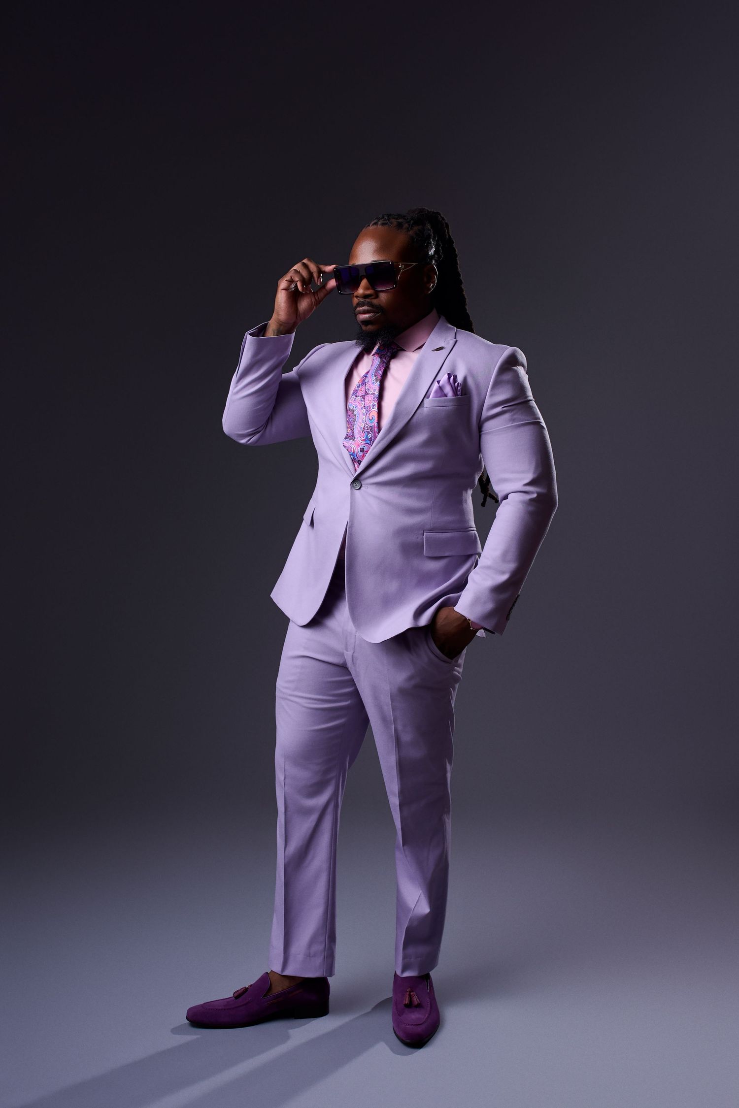 Stylish man in lavender suit with matching shoes, floral tie, and sunglasses posing against gray background.