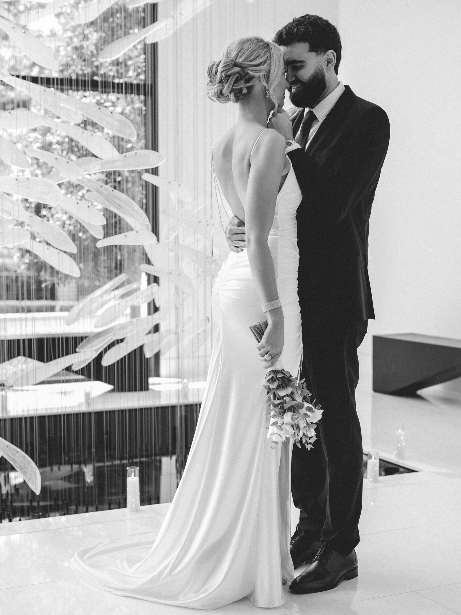 Black and white wedding photo of bride in backless gown and groom in suit sharing an intimate moment indoors. Now booking 2027 weddings