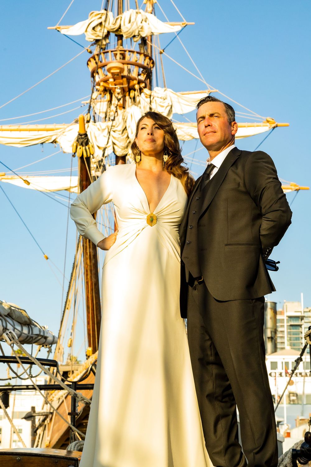 Couple in formal attire poses before tall ship at golden hour.