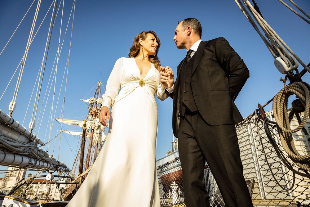 Bride and groom share a romantic moment on a tall ship deck, dressed in elegant wedding attire under a clear blue sky.