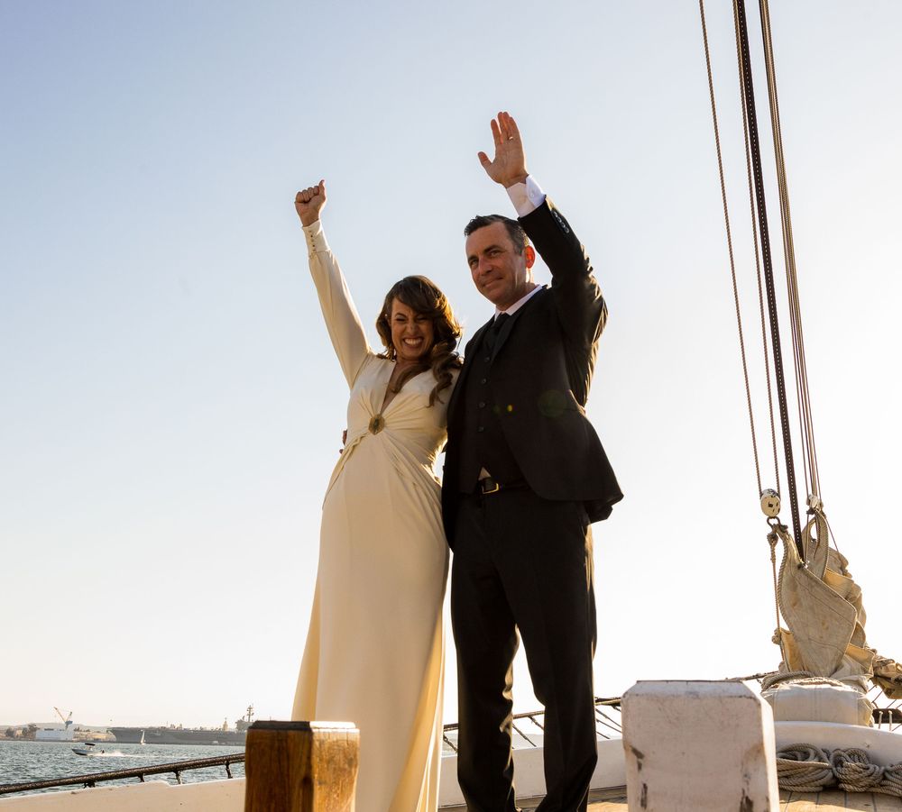 Joyful couple celebrating on a sailboat deck, woman in white gown and man in dark suit, both raising arms.