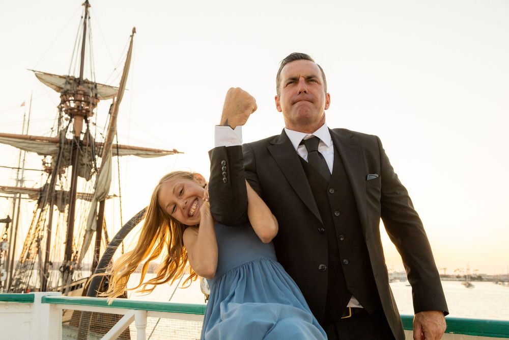 Man in suit and woman in blue dress pose dramatically on ship deck with tall sailing vessel in background.