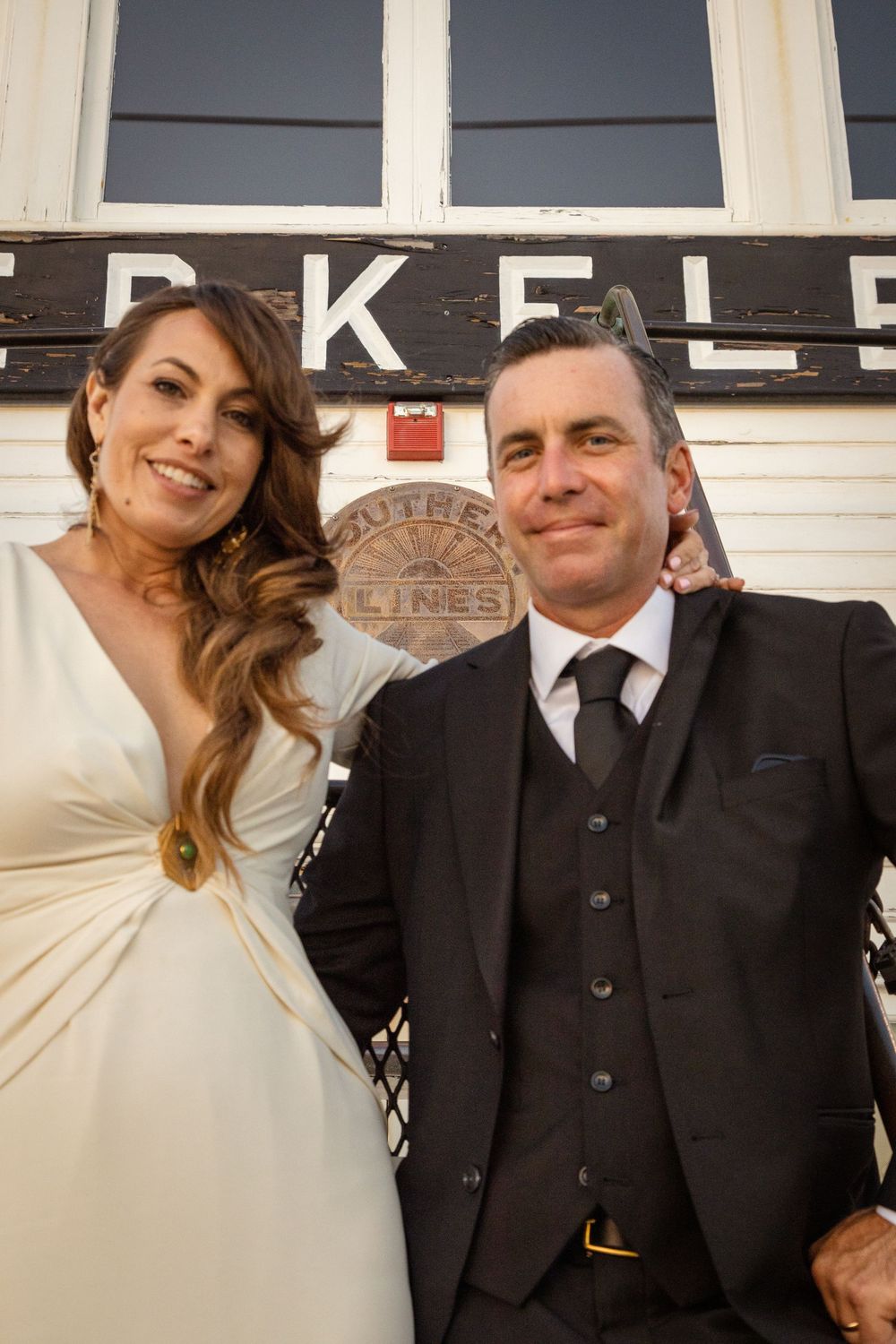 Couple in formal attire posing in front of Berkeley Southern Lines sign, woman in white dress and man in dark suit.