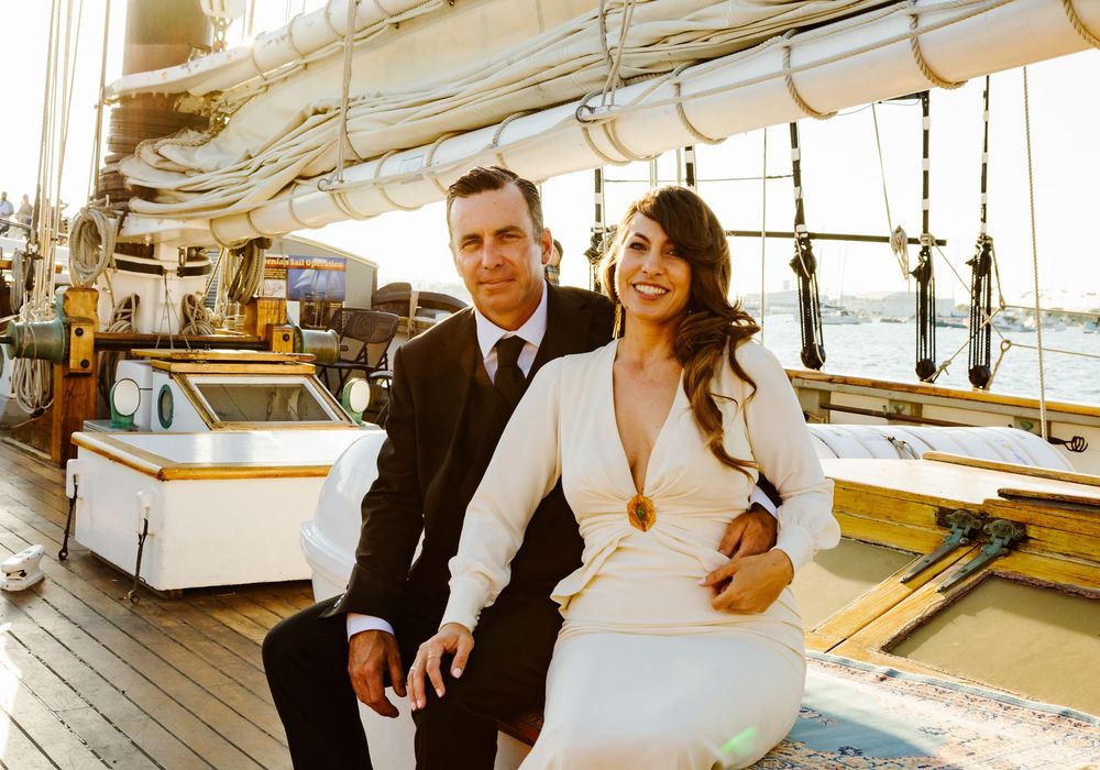 A couple in wedding attire poses on a sailboat deck, bride in white gown and groom in black suit.
